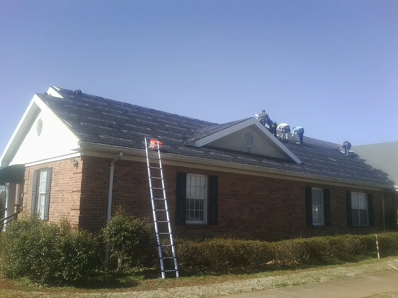 Crew tearing shingles off a brick ranch roof with a ladder set — Metal Roof Repair work by our crew in Judsonia