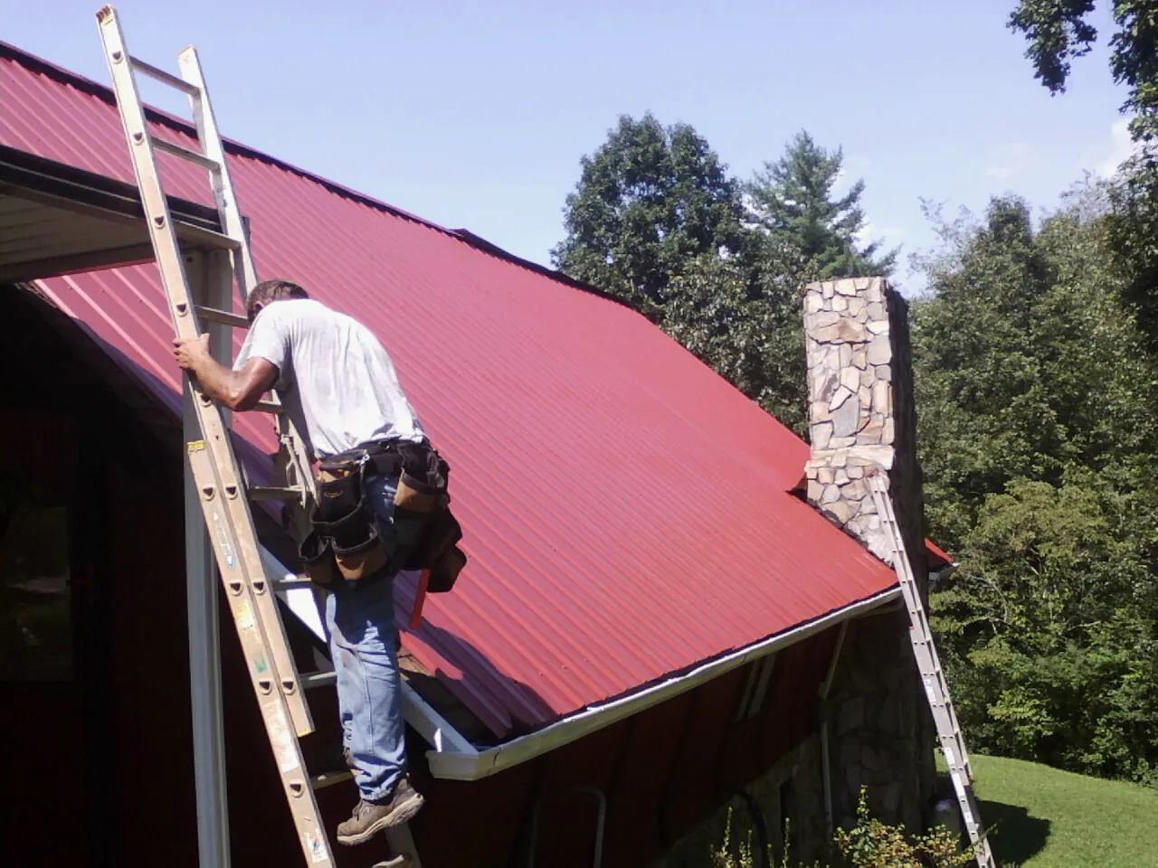 Roofer on a ladder beside a red metal roof and stone chimney — Soffit & Fascia Repair by Roofing Brothers in Judsonia